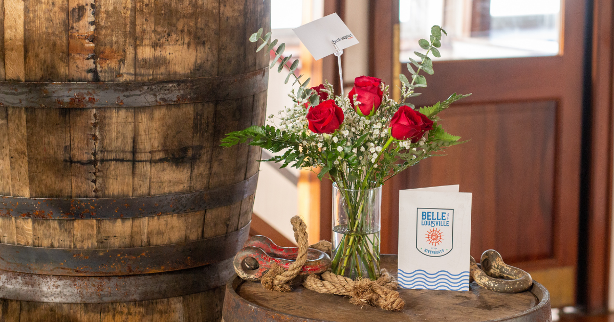A half dozen roses in a vase with foliage. On the right is a card with the Belle of Louisville Riverboats logo. Both are on top of a barrel with rope. On the left are two more barrels stacked on top of each other. In the background are windows and a brown wooden door.