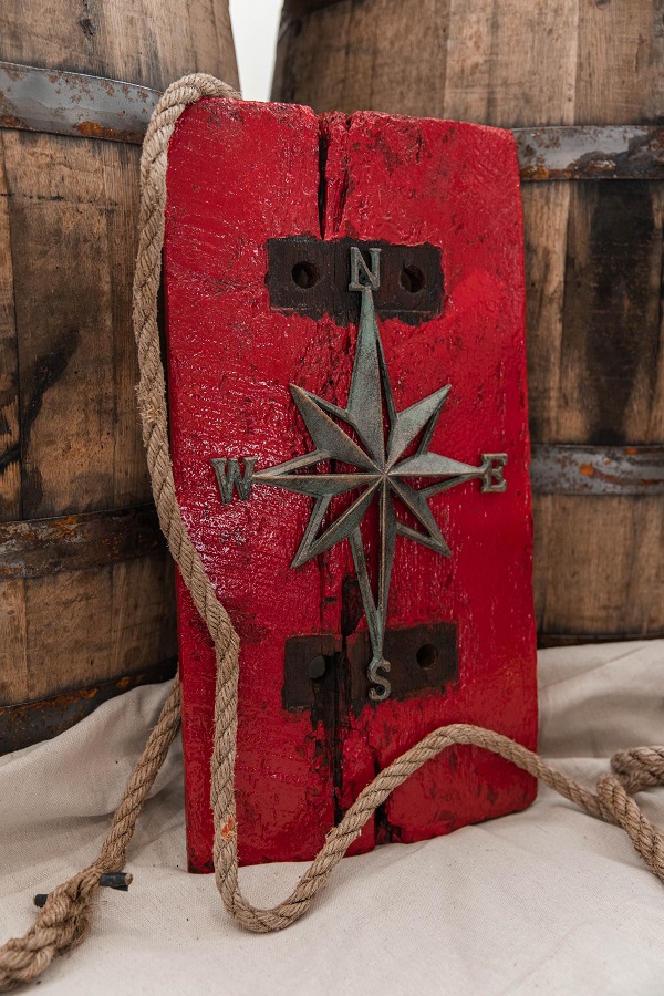 A red piece of paddlewheel wood art with a compass rose on it leaning up against two bourbon barrels. A decorative rope is draped on it.