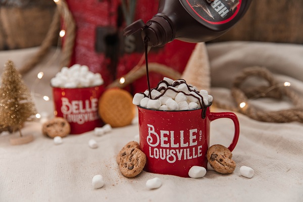 Two red mugs with the Belle of Louisville logo, one in the background and one in the foreground. Both have marshmallows in the mug and cookies resting against the side of the mug. The mug in the foreground has chocolate syrup being drizzled on it. 