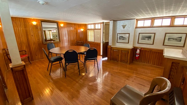 A room with a wooden floor with a back wall of wood paneling. The wall on the right is partially wood paneled and is lined with framed photos on the wall with windows towards the ceiling. In the back center of the room is a table with chairs.