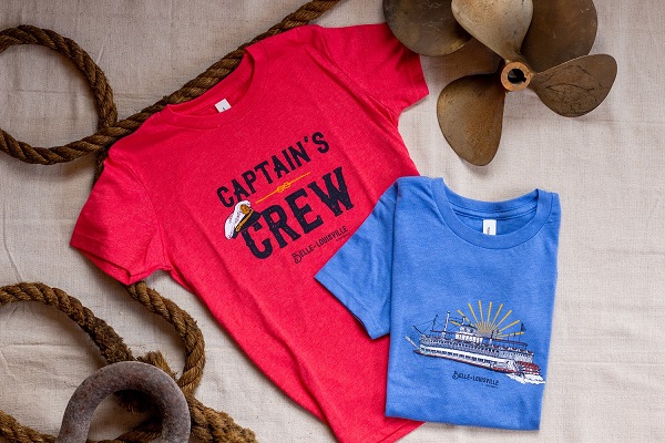 A flat lay of two youth t-shirts. A red shirt with the text Captain's Crew on it is laid flat. On top of it in the corner is a folded blue shirt with a graphic of the Belle of Louisville on it.